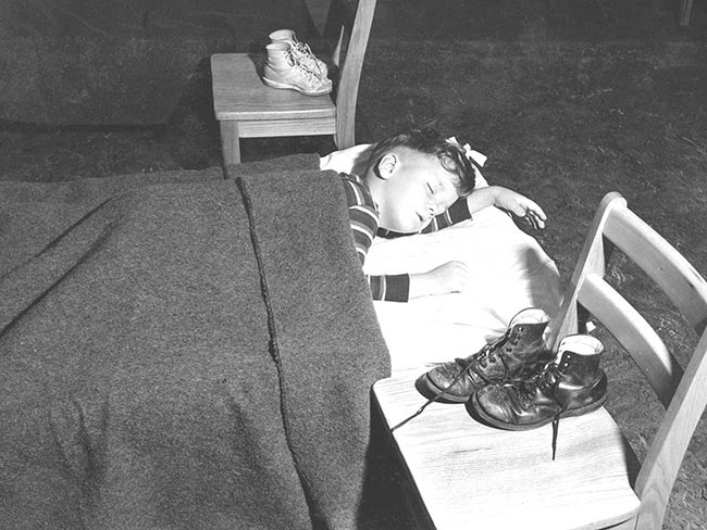 Black and white image of a young boy sleeping in a cot with his boots on a chair next to him. 