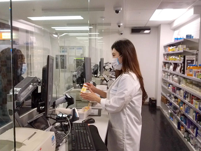 Pharmacist in face mask hands a member her prescription from behind plexiglass.